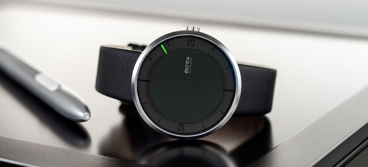Die Abbildung zeigt die BOTTA NOVA Automatik, eine minimalistische Einzeigeruhr mit schwarzem Zifferblatt und dünnem Titangehäuse. Der markante grüne Stundenindikator am Rand ersetzt klassische Zeiger und macht die Uhr zu einer außergewöhnlichen Designuhr. Auf einem Schreibtisch mit Stift inszeniert, unterstreicht das Bild den modernen Charakter dieser hochwertigen Automatikuhr, die Sie online kaufen oder bestellen können.