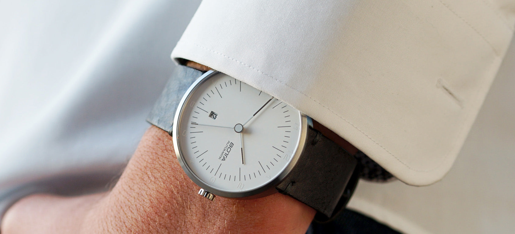 Abgebildet ist die TRES Automatik von BOTTA Design, getragen zu einem weißen Hemd. Diese elegante Automatikuhr kombiniert ein aufgeräumtes weißes Zifferblatt und Datum mit einem 44 mm Titangehäuse und schwarzem Lederarmband. Als mehrfach ausgezeichnete Designuhr vereint sie reduziertes Bauhaus-Design mit Präzision und Langlebigkeit. Eine exklusive Armbanduhr für Individualisten, die eine hochwertige Uhr kaufen oder bestellen möchten.