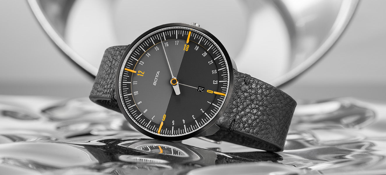 Die BOTTA UNO 24 Quarzuhr 45 mm Black Edition mit markanten gelben Markierungen bei 6, 12, 18 und 24 Uhr, präsentiert auf reflektierendem Untergrund. Das dunkle 24-Stunden-Zifferblatt mit nur einem Zeiger vermittelt Klarheit und Ruhe im Umgang mit der Zeit. Schwarzes Lederarmband und reduziertes Design unterstreichen den Anspruch als preisgekrönte Designuhr. Eine exklusive Einzeigeruhr – jetzt kaufen oder bestellen.