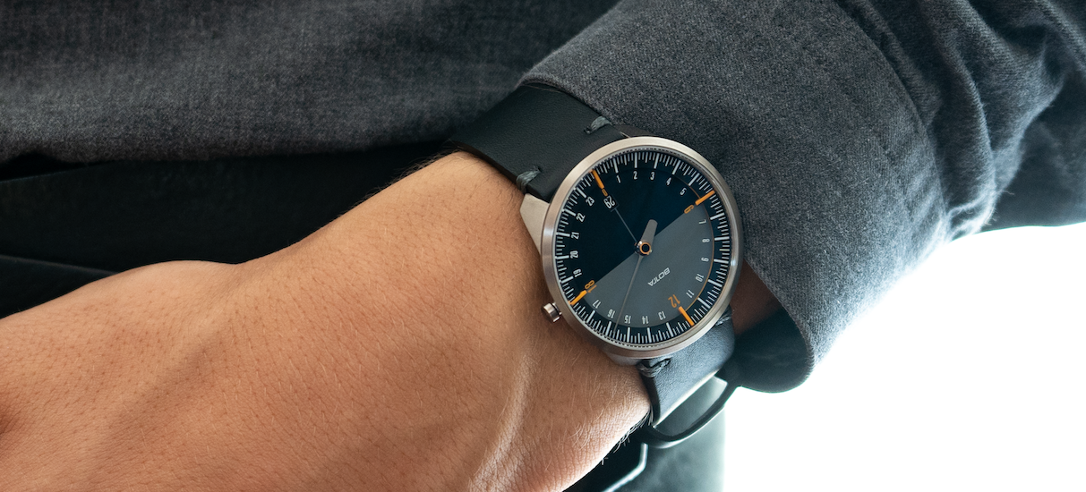 Die BOTTA UNO 24 Quarzuhr 40 mm mit schwarzem Lederarmband am Handgelenk getragen. Das Zifferblatt zeigt die charakteristische 24-Stunden-Einteilung mit einem einzigen Zeiger. Dezente orangefarbene Markierungen bei 6 und 12 Uhr setzen moderne Akzente und heben die minimalistische Gestaltung hervor. Diese preisgekrönte Designuhr vermittelt Gelassenheit und Zeitbewusstsein. Eine stilvolle Einzeigeruhr – jetzt bestellen oder kaufen.