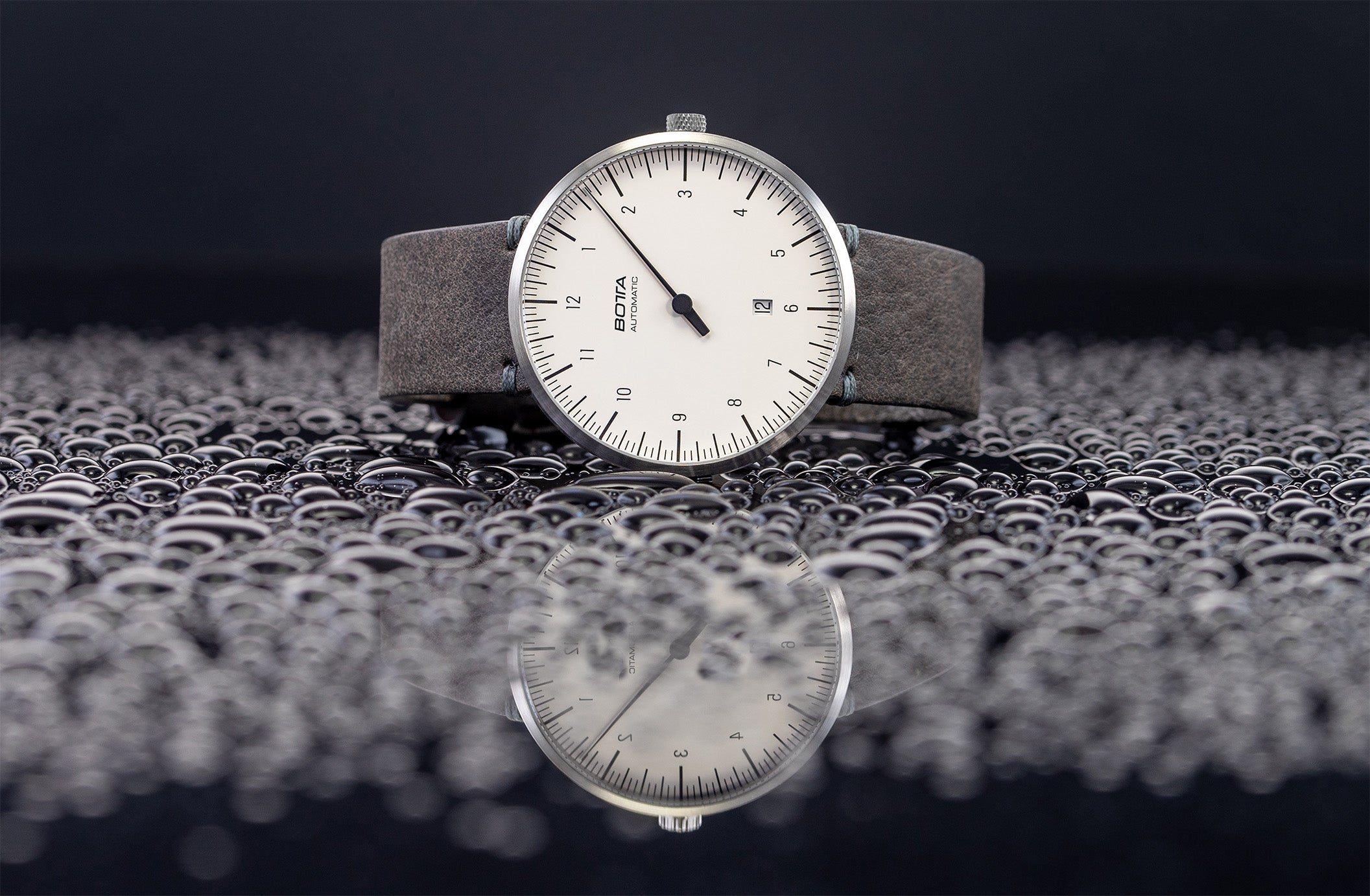 Die Einzeigeruhr liegt auf einer von Wassertropfen bedeckten Fläche, wodurch ihre Widerstandsfähigkeit und Alltagstauglichkeit hervorgehoben werden. Das weiße Zifferblatt mit nur einem eleganten schwarzen Zeiger und klarer Skala steht für Minimalismus und intuitive Zeitablesung. Technisch überzeugt die Automatikuhr mit dem präzisen Uhrwerk, kratzfestem Saphirglas und 5 ATM Wasserdichtigkeit. Wer eine außergewöhnliche Armbanduhr oder eine Uhr mit einem Zeiger kaufen möchte, findet hier ein Original.