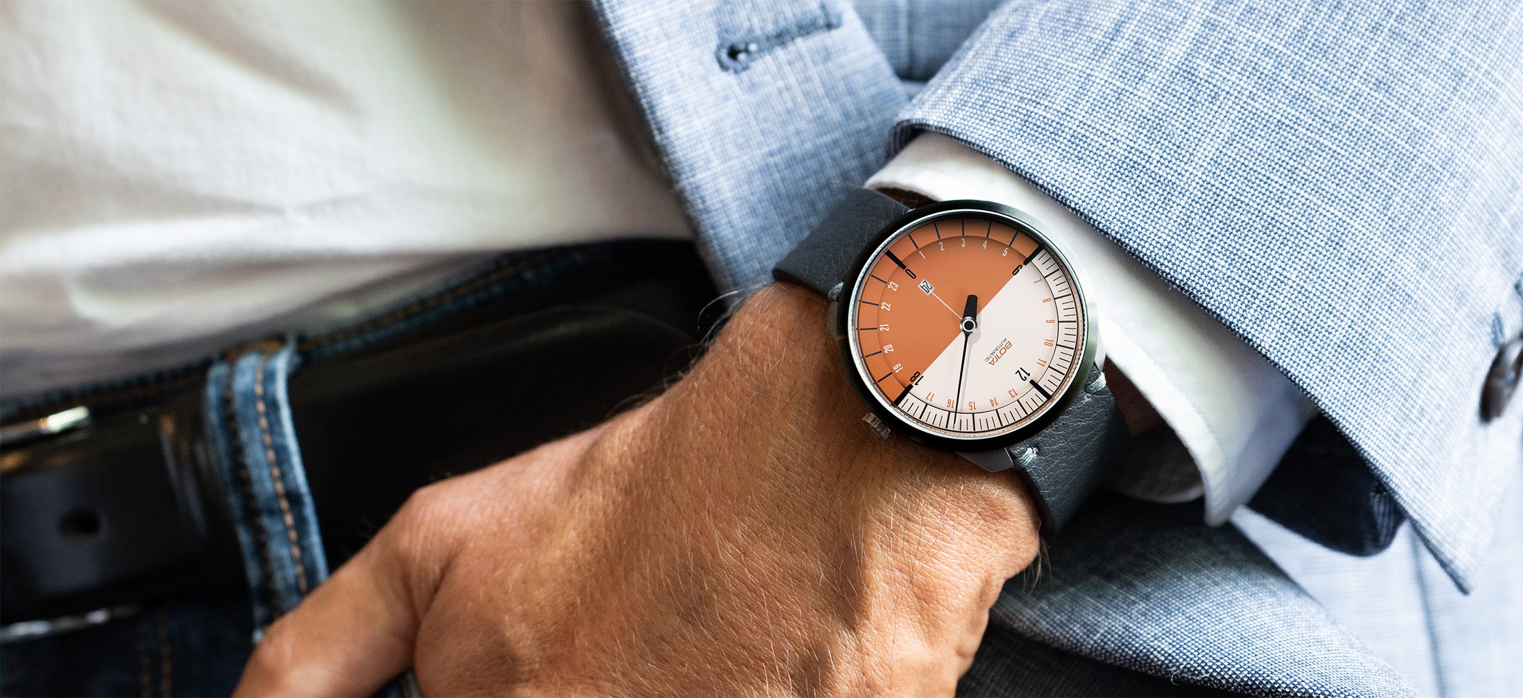 Ein Mann trägt die BOTTA UNO 24 Your Time am Handgelenk, kombiniert mit elegantem Sakko und Jeans. Die hochwertige Automatikuhr zeigt ihr zweifarbiges Terracotta-weißes Zifferblatt, das den Tagesverlauf in 24 Stunden mit nur einem Zeiger darstellt. Diese Einzeiger Automatikuhr verbindet minimalistisches Design mit Präzision und Stil. Eine 24-Stunden Uhr, die Klarheit und Individualität ausstrahlt. Jetzt kaufen oder bestellen.