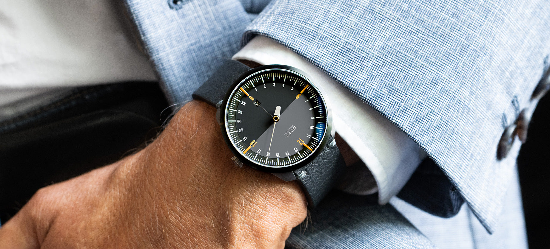 Das Bild zeigt die BOTTA UNO 24 Automatik am Handgelenk, getragen zu einem eleganten Anzug. Das schwarze 24-Stunden-Zifferblatt mit gelben Akzenten und nur einem Zeiger verdeutlicht das Einzeigerprinzip dieser Automatikuhr. Sie zeigt den gesamten Tagesverlauf in einer Umdrehung. Mit Tri-Titan-Gehäuse, doppelt gewölbtem Saphirglas und dem präzisen Sellita SW330 Automatikwerk ist sie eine hochwertige Designuhr für Anspruchsvolle, die eine exklusive Armbanduhr mit einem Zeiger kaufen oder bestellen möchten.