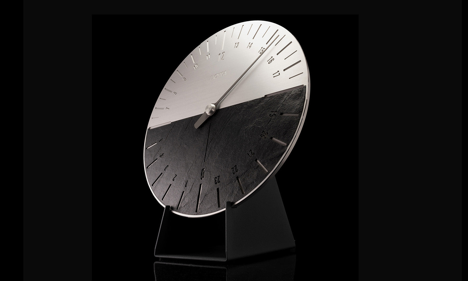 Die UNO 24 Slate Wanduhr von BOTTA im seitlichen Blick auf schwarzem Hintergrund: Das 24-Stunden-Zifferblatt zeigt eine elegante Zweiteilung – obere Hälfte aus gebürstetem Edelstahl, untere Hälfte aus natürlichem Schiefer. Ein einzelner Zeiger symbolisiert die Einzeigerphilosophie, die den Tag in einem einzigen Umlauf erfasst. Diese Designwanduhr mit Schiefer vereint Minimalismus und Naturmaterialien. Jetzt exklusiv UNO 24 Wanduhr kaufen oder bestellen