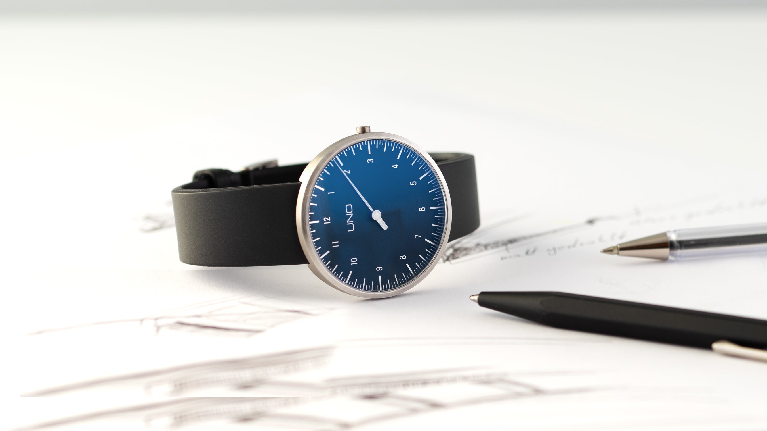 Die BOTTA UNO Einzeigeruhr Titan Quarzuhr zeigt sich hier mit tiefblauem Zifferblatt, einem einzelnen weißen Zeiger und minimalistischer Minuterie. Das Gehäuse aus Titan unterstreicht den leichten, puristischen Charakter dieser Designuhr. Im Bild liegt die Einzeigeruhr neben Skizzen und Schreibgeräten – ein Symbol für Kreativität und Präzision. Eine außergewöhnliche Armbanduhr für Menschen, die Zeit bewusst erleben möchten. Jetzt online kaufen oder bestellen.