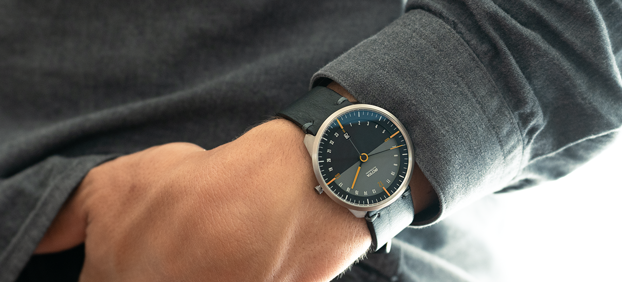 Auf dem Bild ist die BOTTA TRES 24 Automatikuhr mit schwarzem Zifferblatt und orangefarbenen Akzenten am Handgelenk zu sehen. Das 24-Stunden-Design mit zusätzlichem Minuten- und Sekundenzeiger ermöglicht ein entspanntes, aber präzises Zeitgefühl. Das Titangehäuse und das schwarze Lederarmband strahlen Modernität und Robustheit aus. Diese hochwertige Designuhr ist eine besondere Automatikuhr mit innovativem Konzept – ideal für alle, die eine exklusive Automatikuhr kaufen oder bestellen möchten.