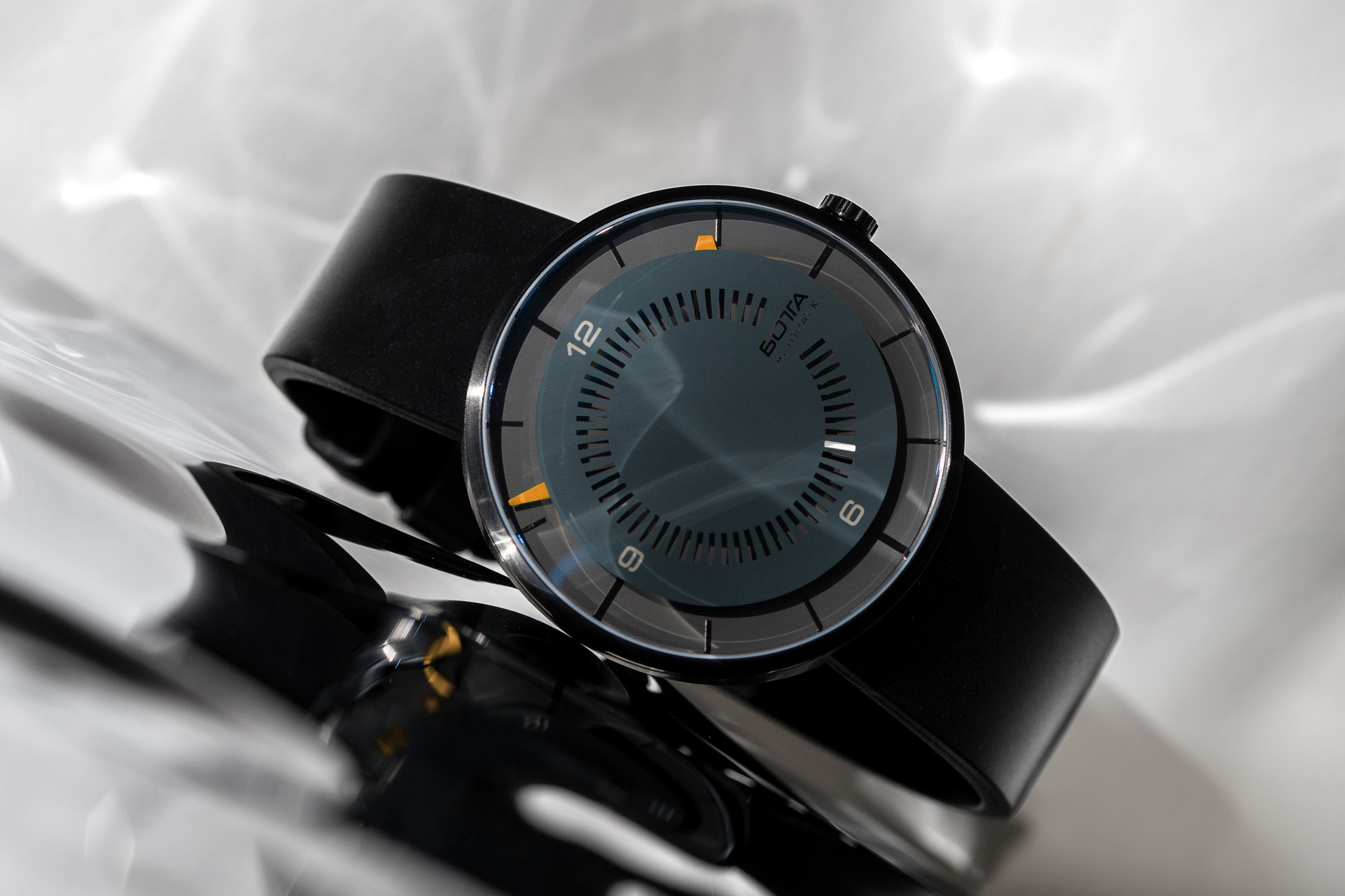 Die BOTTA CLAVIUS Automatik 44 mm präsentiert sich in einem künstlerischen Setting mit silbrig schimmerndem Hintergrund. Die Designuhr mit futuristischem Prinzip zeigt ein tiefschwarzes Zifferblatt, markante orange Akzente und minimalistisches Industriedesign. Eine Automatikuhr für Individualisten, die eine außergewöhnliche Armbanduhr mit besonderem Look  kaufen oder bestellen möchten – modern, präzise und einzigartig.