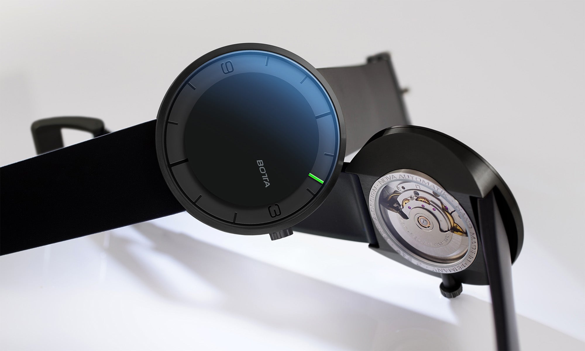 Auf dem Bild ist die BOTTA NOVA Automatik in der Farbe Schwarz zu sehen – eine außergewöhnlich flache Automatikuhr und puristische Designuhr. Das äußerst flache Titangehäuse mit tiefschwarzem Zifferblatt unterstreicht das radikal minimalistische Anzeigeprinzip: nur ein einziger, feiner Zeiger weist auf die Stunden. Die Rückseite offenbart das präzise Sellita SW200 Automatikwerk durch den Glasboden, ein Symbol für Schweizer Präzision und deutsche Gestaltungskunst. 