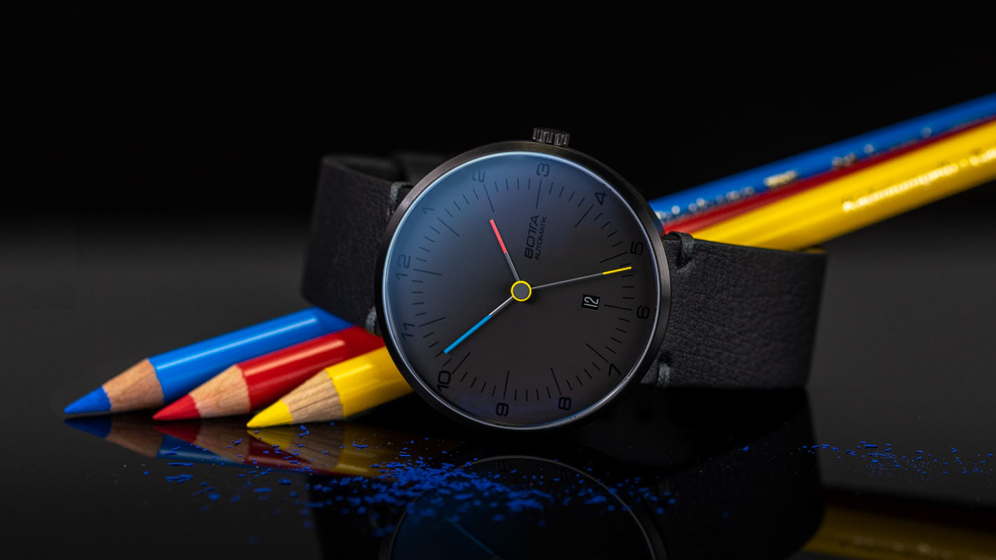 Das Bild zeigt die BOTTA TRES colores Automatik in einer künstlerischen Inszenierung mit roten, blauen und gelben Buntstiften. Das schwarze Zifferblatt und die klaren Bauhaus-Linien werden durch die farbigen Zeiger hervorgehoben. Das 44 mm Edelstahlgehäuse ist DLC-beschichtet, kratzfestes Saphirglas schützt das Zifferblatt, im Inneren arbeitet ein Sellita SW200-1 Automatikwerk. Eine preisgekrönte Designuhr und Automatikuhr – ideal für Individualisten, die eine besondere Design-Armbanduhr suchen.